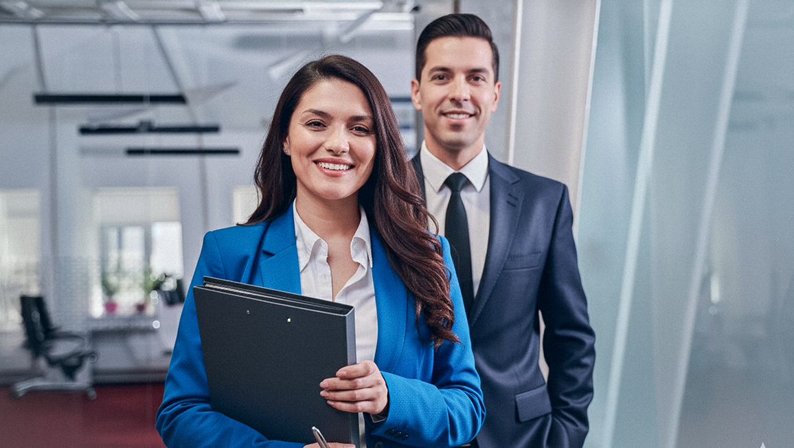 Dos profesionales de negocios, una mujer con traje azul y un hombre con traje oscuro, sonriendo en una oficina.